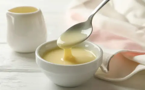 Latte condensato denso e cremoso, perfetto per dolci fatti col Bimby, cola da un cucchiaio in una ciotola bianca.