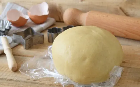 Impasto di pasta frolla con olio, pronto per essere trasformato in biscotti con stampini a forma di stella e fiore. Guscio d'uovo e mattarello completano la scena.