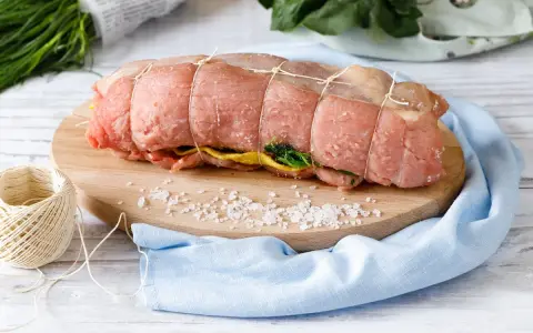 Un arrosto al forno pronto per essere cucinato, legato con spago, su un tagliere con sale grosso e verdure.