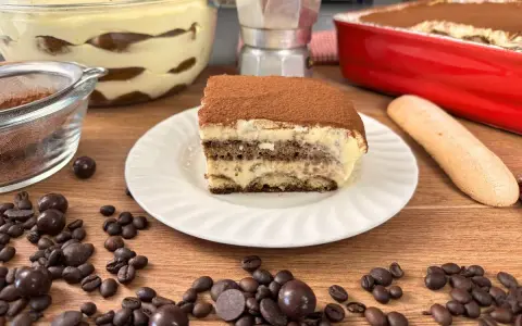 Una fetta di tiramisù con uova pastorizzate, cosparso di cacao, su un piatto bianco, circondato da chicchi di caffè e cioccolatini.