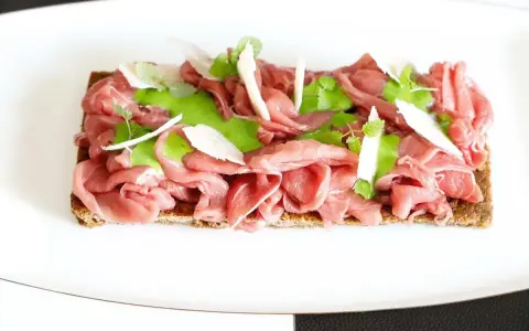 Delicato carpaccio di manzo su crostino integrale, guarnito con crema verde, scaglie di parmigiano e foglioline fresche.