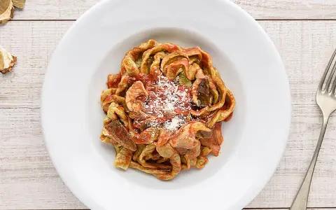 Un piatto di tagliatelle ai funghi, condite con sugo rosso e parmigiano grattugiato, su un tavolo di legno.