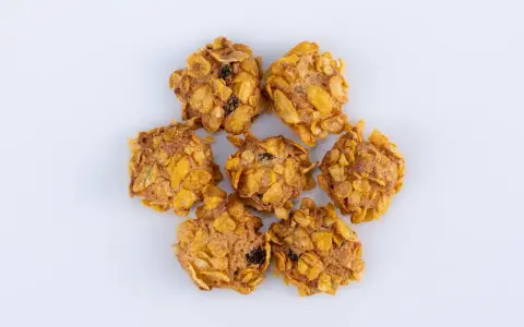 Biscotti croccanti, simili a rose del deserto, con fiocchi di mais e uvetta, disposti a cerchio su sfondo bianco.