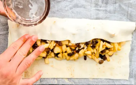 Mani che preparano uno strudel di mele con pasta sfoglia, riempiendolo con mele a cubetti, uvetta e pinoli.