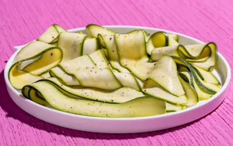 Fettine sottili di zucchine marinate crude, condite con pepe nero, su un piatto bianco, su un tavolo rosa acceso.