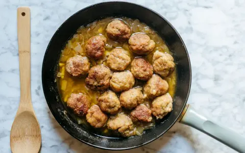 Polpette dorate in padella con cipolle, pronte per essere gustate. Un cucchiaio di legno attende accanto.