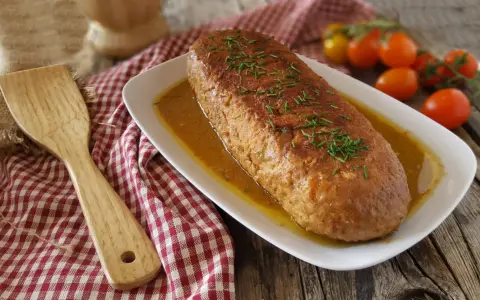 Polpettone dorato in padella, guarnito con erba cipollina, su un piatto bianco con salsa, accanto a pomodorini e una spatola di legno.