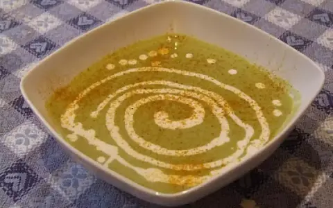 Una ciotola quadrata bianca contiene una vellutata crema di piselli, decorata con un vortice di panna e una spolverata di spezie.