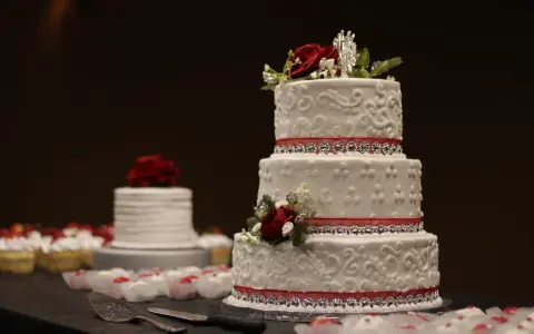 Torta nuziale a tre piani decorata con pasta di zucchero, rose rosse e nastri scintillanti. Cupcake e un'altra torta sullo sfondo.