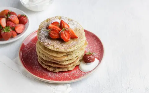 Pila di pancake senza glutine guarniti con fragole fresche e zucchero a velo, serviti su un piatto rosso con un cucchiaio di panna.