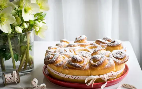 Una deliziosa torta di rose, cosparsa di zucchero a velo, su un piatto rosso, con un nastro a cuoricini. Accanto, un vaso con fiori bianchi.