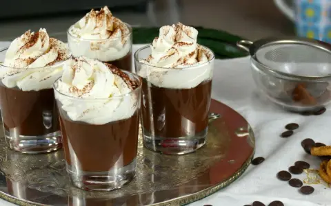 Quattro bicchierini di budino al cioccolato, guarniti con panna montata e cacao in polvere, su un vassoio d'argento.