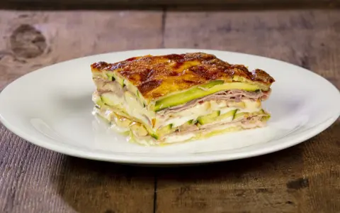 Una fetta di parmigiana di zucchine bianca, stratificata con prosciutto e formaggio filante, su un piatto bianco.