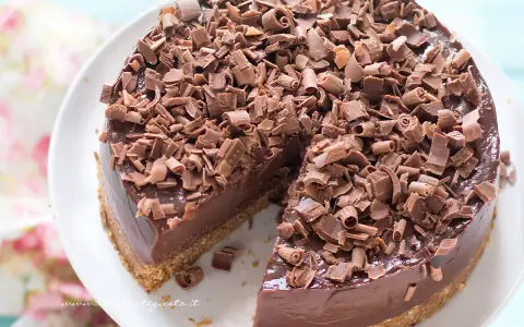 Cheesecake alla Nutella golosa, ricoperta di riccioli di cioccolato fondente. Una fetta è già stata servita, invitando a gustarla.