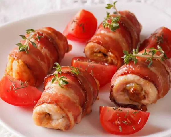 Involtini di pollo avvolti in pancetta croccante, guarniti con timo fresco e serviti con spicchi di pomodoro.