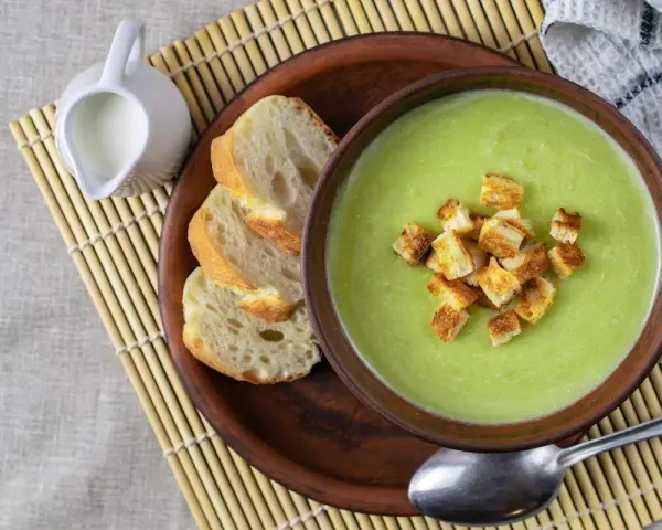 Una ciotola di cremosa crema di zucchine con crostini dorati, accompagnata da pane fresco e una caraffa di latte.