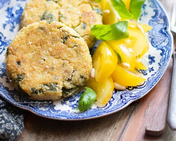 Hamburger di zucchine dorati e croccanti, serviti con pomodorini gialli e basilico fresco su un piatto blu e bianco.