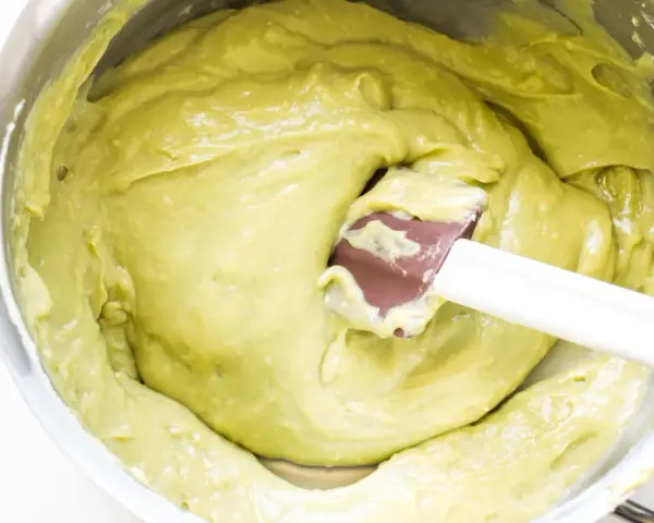 A spatula mixes a smooth, pale green pistachio cream in a stainless steel bowl.