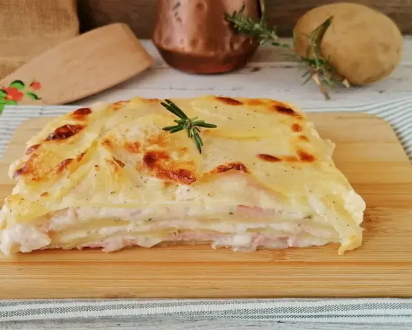 Una fetta di parmigiana di patate dorata e filante, con strati di patate, prosciutto e besciamella, guarnita con rosmarino.