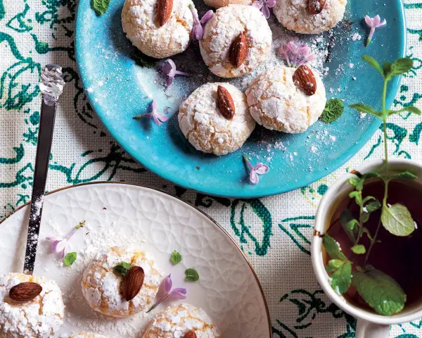 Biscotti al cocco spolverati di zucchero a velo, decorati con mandorle e fiori viola, accanto a una tazza di tè alla menta.