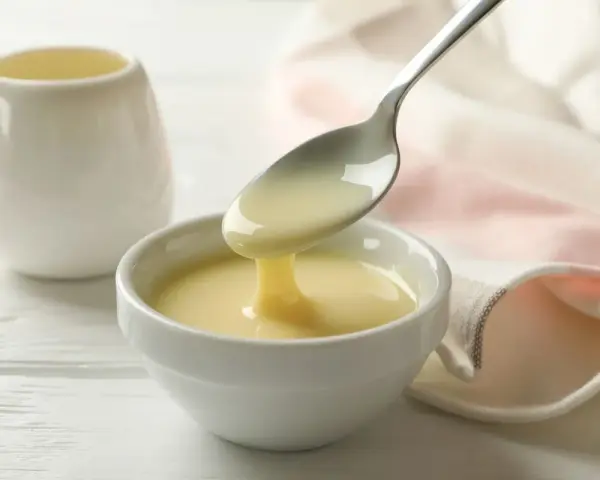 Latte condensato denso e cremoso, perfetto per dolci fatti col Bimby, cola da un cucchiaio in una ciotola bianca.