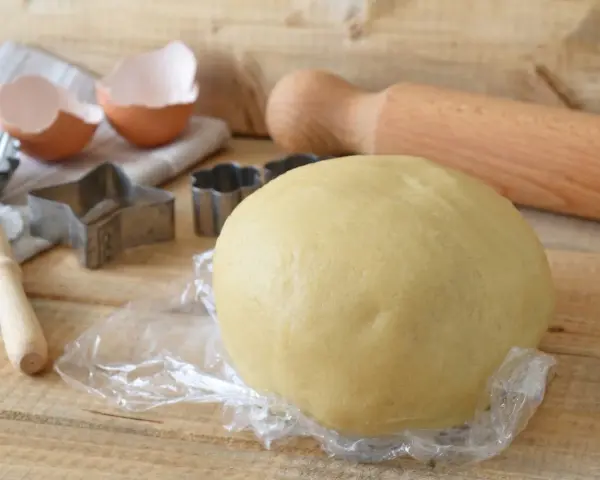 Impasto di pasta frolla con olio, pronto per essere trasformato in biscotti con stampini a forma di stella e fiore. Guscio d'uovo e mattarello completano la scena.