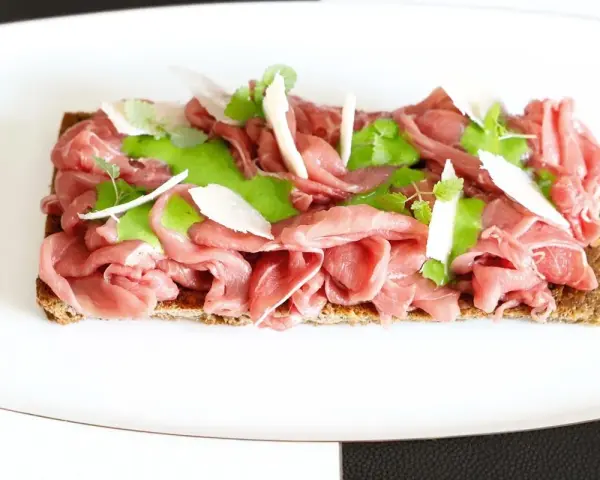 Delicato carpaccio di manzo su crostino integrale, guarnito con crema verde, scaglie di parmigiano e foglioline fresche.