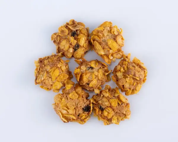 Biscotti croccanti, simili a rose del deserto, con fiocchi di mais e uvetta, disposti a cerchio su sfondo bianco.
