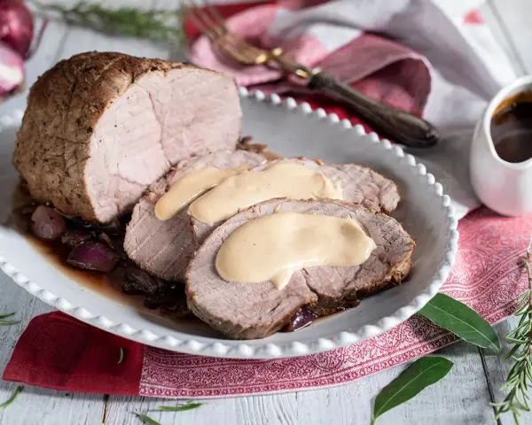 Arrosto di vitello morbido con sughetto benedetta, servito su un piatto bianco con cipolle rosse e rosmarino.