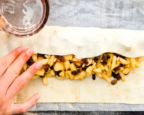 Mani che preparano uno strudel di mele con pasta sfoglia, riempiendolo con mele a cubetti, uvetta e pinoli.