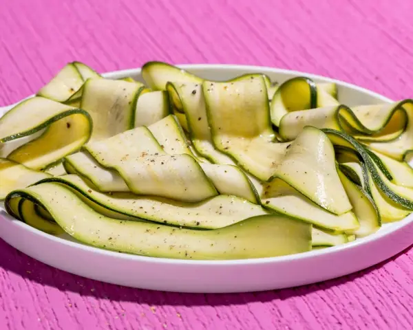 Fettine sottili di zucchine marinate crude, condite con pepe nero, su un piatto bianco, su un tavolo rosa acceso.