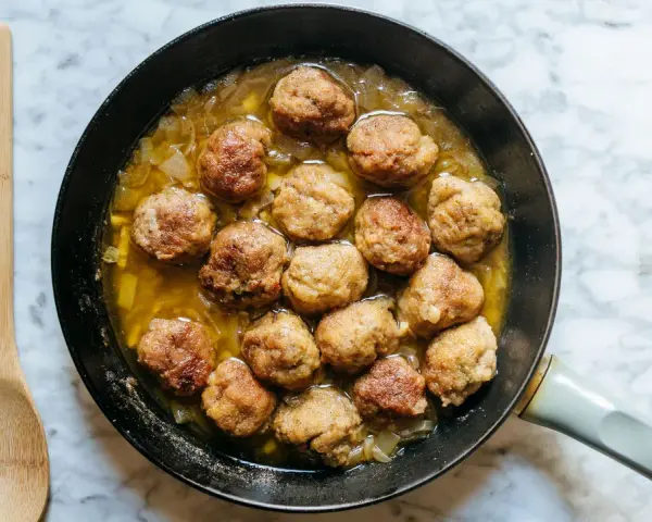 Polpette dorate in padella con cipolle, pronte per essere gustate. Un cucchiaio di legno attende accanto.