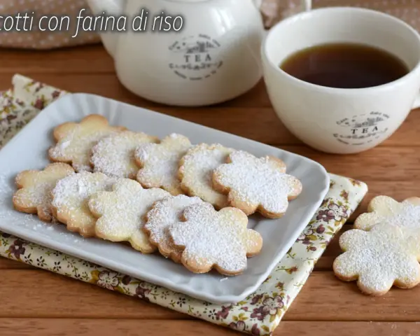Biscotti con farina di riso a forma di fiore, spolverati di zucchero a velo, accanto a una tazza di tè e una teiera.
