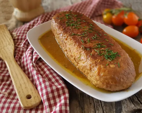 Polpettone dorato in padella, guarnito con erba cipollina, su un piatto bianco con salsa, accanto a pomodorini e una spatola di legno.