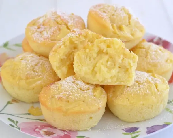 Morbidi muffin alle mele, spolverati di zucchero a velo, su un piatto fiorito. Uno è tagliato a metà, mostrando la soffice mollica.
