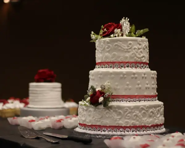 Torta nuziale a tre piani decorata con pasta di zucchero, rose rosse e nastri scintillanti. Cupcake e un'altra torta sullo sfondo.