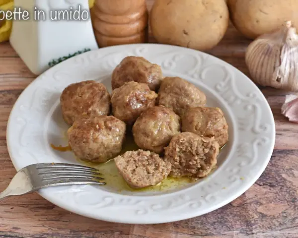 Piatto di polpette in bianco, morbide e succulente, pronte per essere gustate. Sullo sfondo patate e aglio.