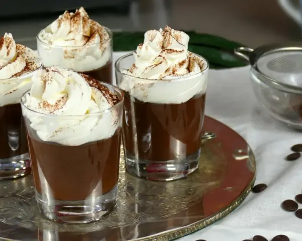 Quattro bicchierini di budino al cioccolato, guarniti con panna montata e cacao in polvere, su un vassoio d'argento.