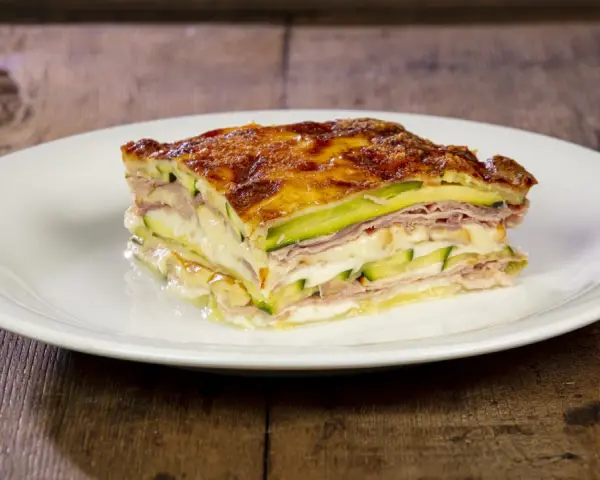 Una fetta di parmigiana di zucchine bianca, stratificata con prosciutto e formaggio filante, su un piatto bianco.