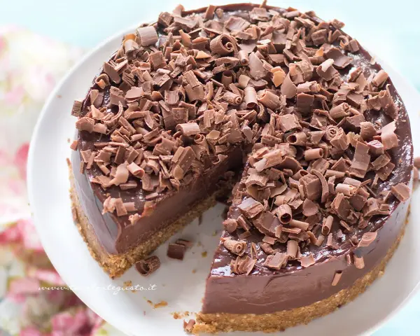 Cheesecake alla Nutella golosa, ricoperta di riccioli di cioccolato fondente. Una fetta è già stata servita, invitando a gustarla.