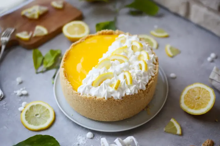 Delizioso cheesecake al limone con panna montata e fettine di limone, circondato da limoni freschi e foglioline verdi.