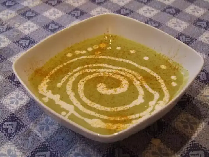 Una ciotola quadrata bianca contiene una vellutata crema di piselli, decorata con un vortice di panna e una spolverata di spezie.