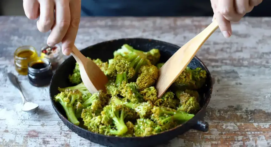 Broccoli in padella, condito con olio e spezie, viene mescolato con due palette di legno.