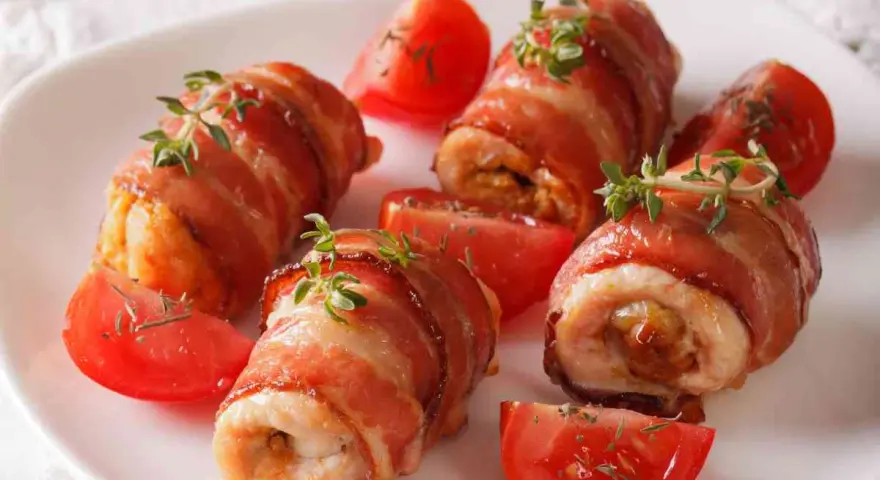 Involtini di pollo avvolti in pancetta croccante, guarniti con timo fresco e serviti con spicchi di pomodoro.