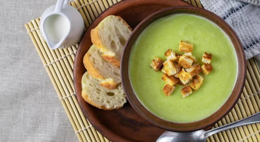 Una ciotola di cremosa crema di zucchine con crostini dorati, accompagnata da pane fresco e una caraffa di latte.
