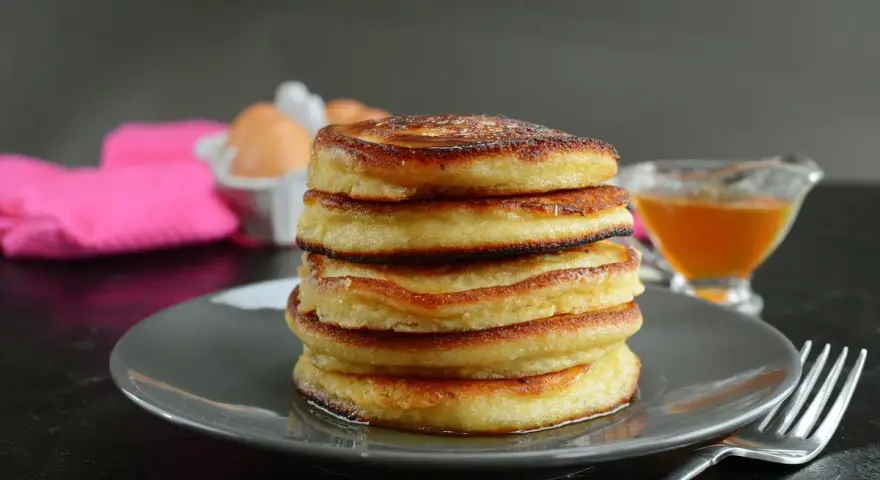 Pila di pancake senza latte, dorati e soffici, con sciroppo d'acero e un tovagliolo rosa sullo sfondo.