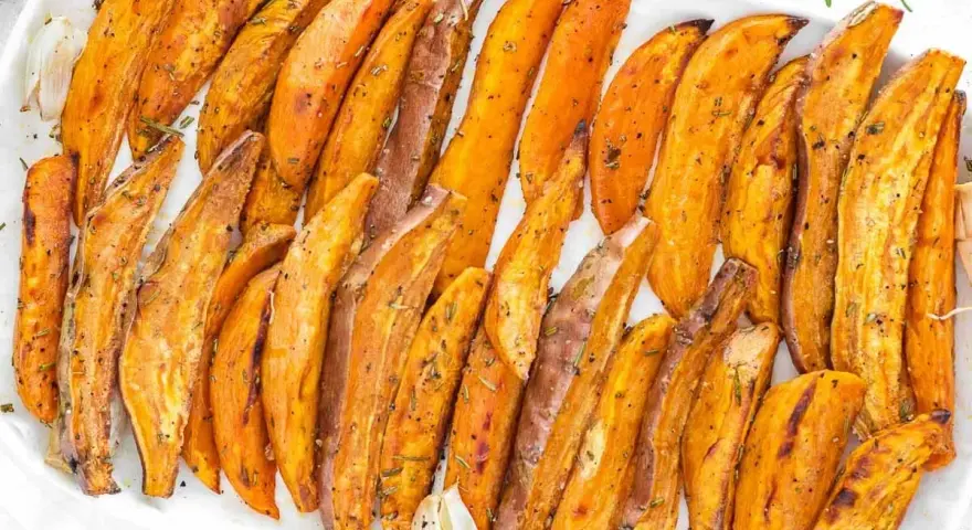 Patate dolci al forno, tagliate a spicchi e condite con erbe aromatiche, servite su un piatto bianco.