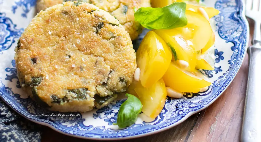Hamburger di zucchine dorati e croccanti, serviti con pomodorini gialli e basilico fresco su un piatto blu e bianco.