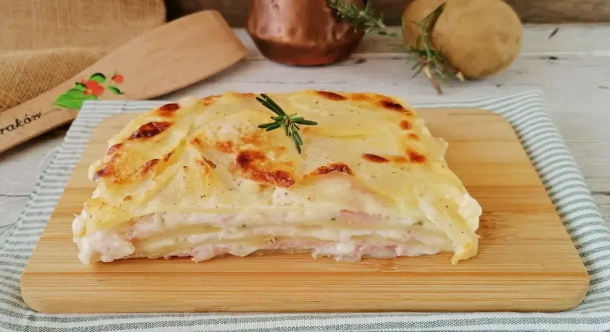Una fetta di parmigiana di patate dorata e filante, con strati di patate, prosciutto e besciamella, guarnita con rosmarino.