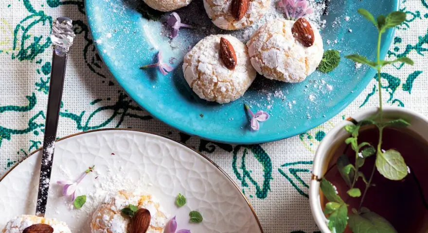 Biscotti al cocco spolverati di zucchero a velo, decorati con mandorle e fiori viola, accanto a una tazza di tè alla menta.